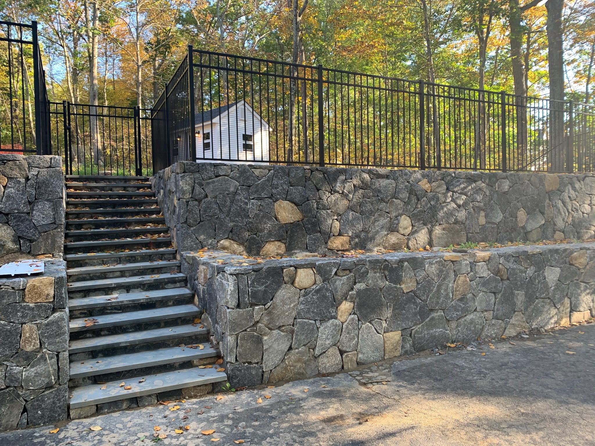 Black aluminum fence with a gate atop a fieldstone retaining wall beside stone steps, autumn trees behind