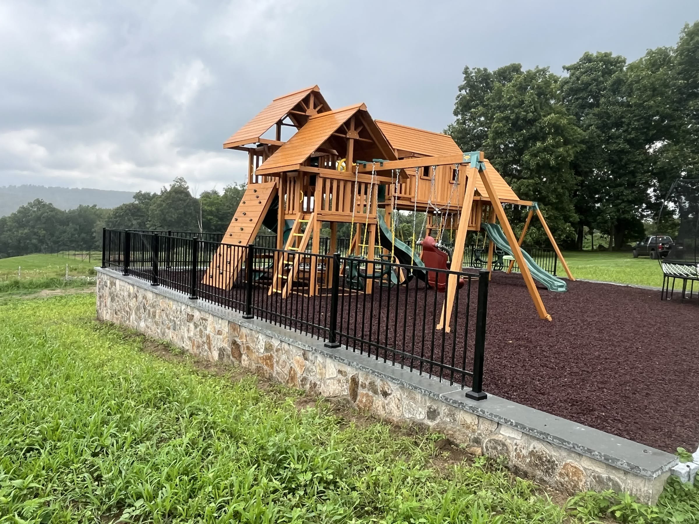 Black aluminum fence atop a stone base wall enclosing a wooden playground set with rubber mulch surfacing in a field