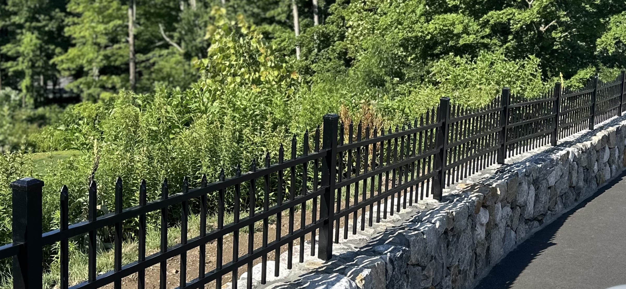 Black aluminum spear-top fence mounted along a mortared stone wall beside a paved driveway with greenery behind