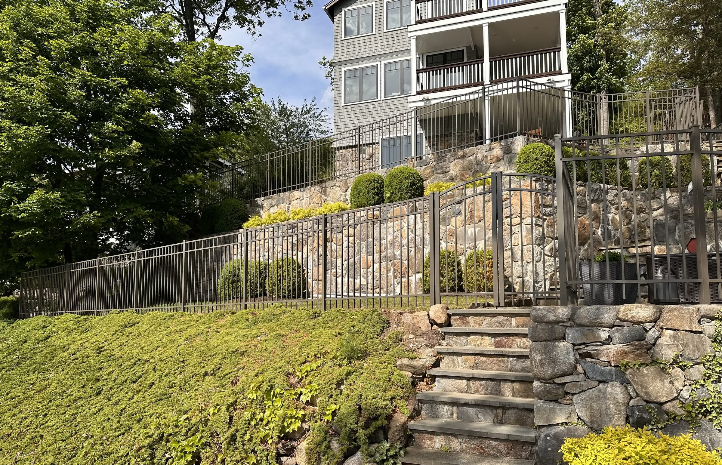 Bronze aluminum fence with an arched walk gate atop a fieldstone retaining wall beside stone steps at a hillside home
