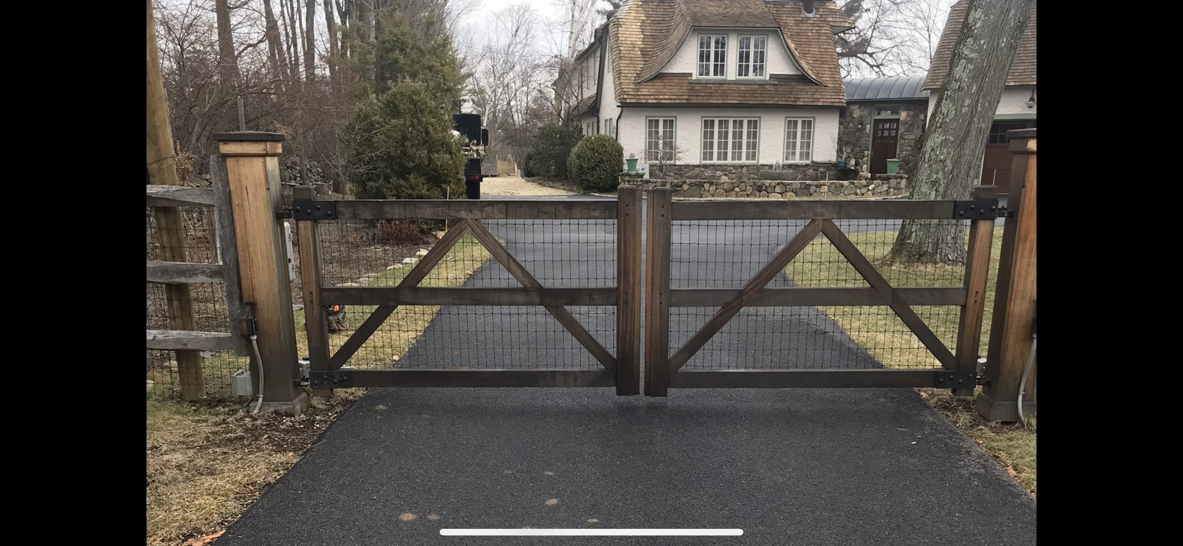 Weathered wood double driveway gate with wire mesh infill and X-bracing on paved drive with house behind