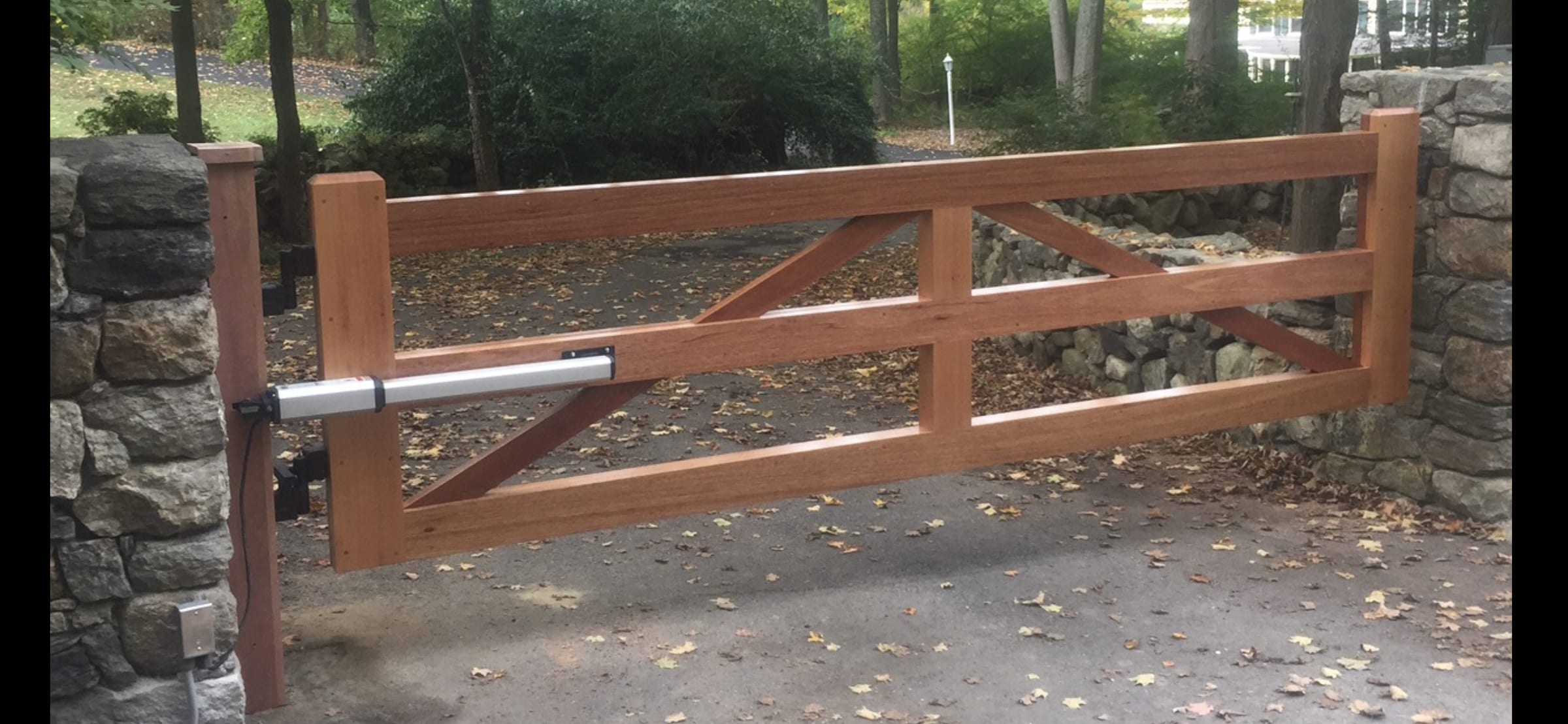 Natural stained wood single-panel driveway gate with automatic opener between stone pillars and stone wall