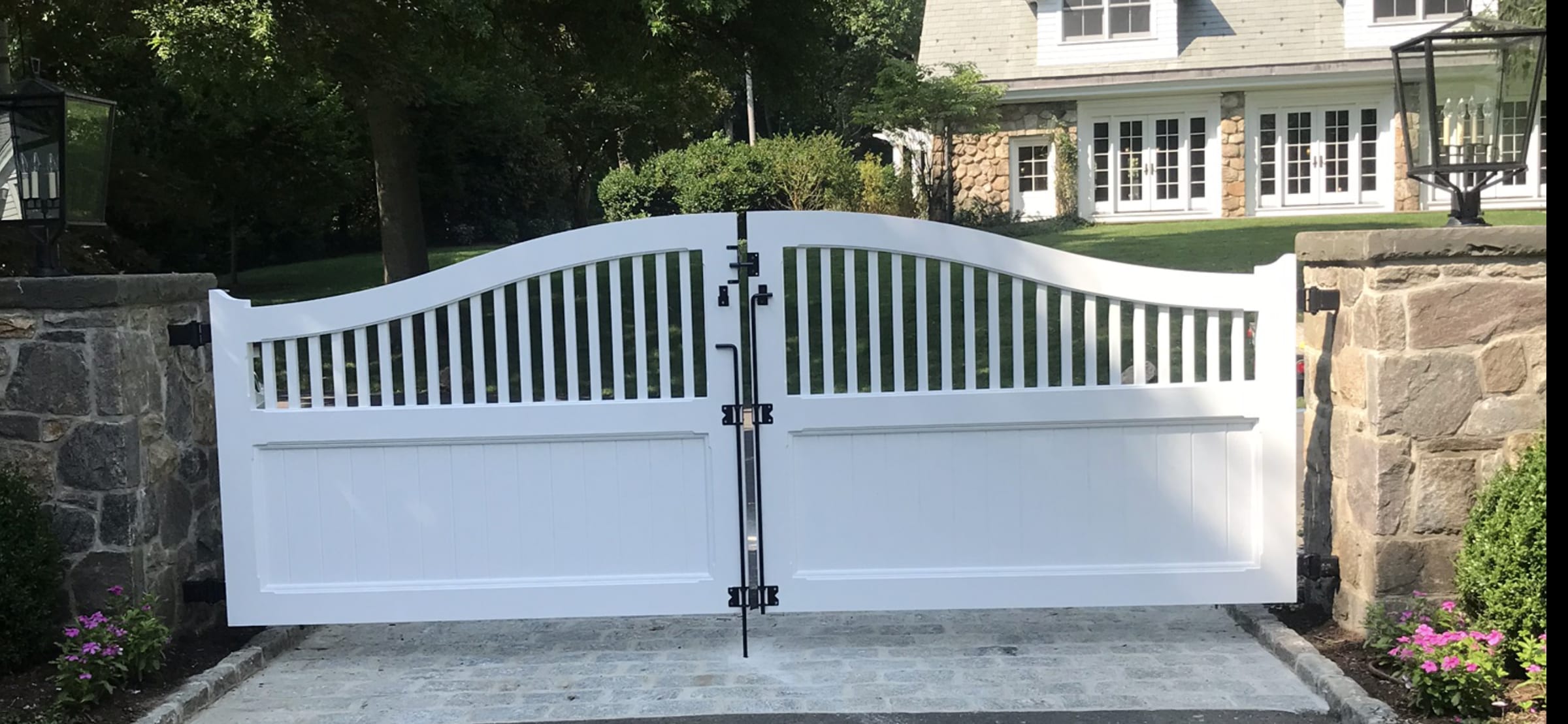 White arched double driveway gate with spindle top and solid lower panels between stone pillars on cobblestone drive