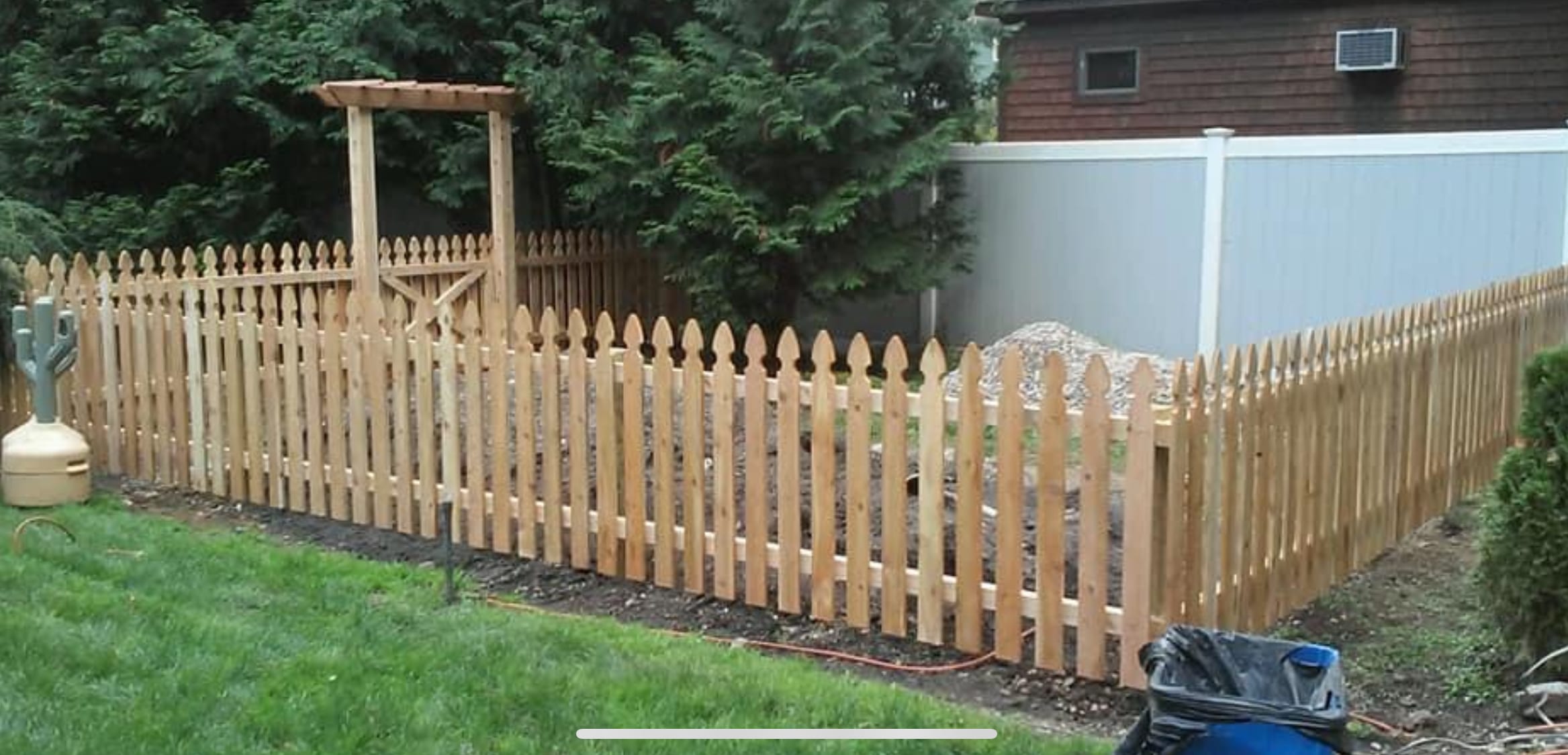New cedar spaced picket fence with pointed French-Gothic tops and an arbor along a backyard garden area.