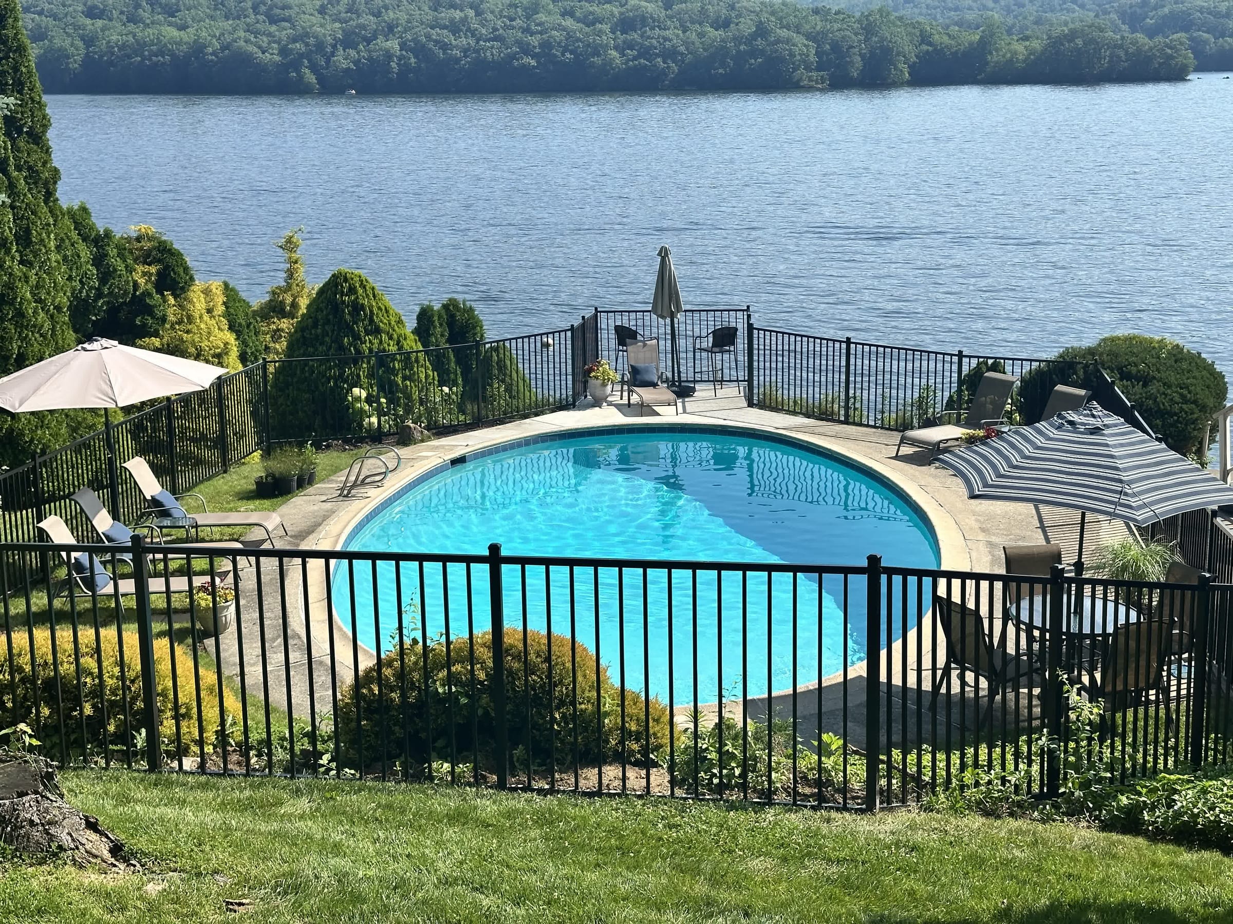 Black aluminum fence enclosing a round inground pool overlooking a lake, with lounge chairs and umbrellas on the deck