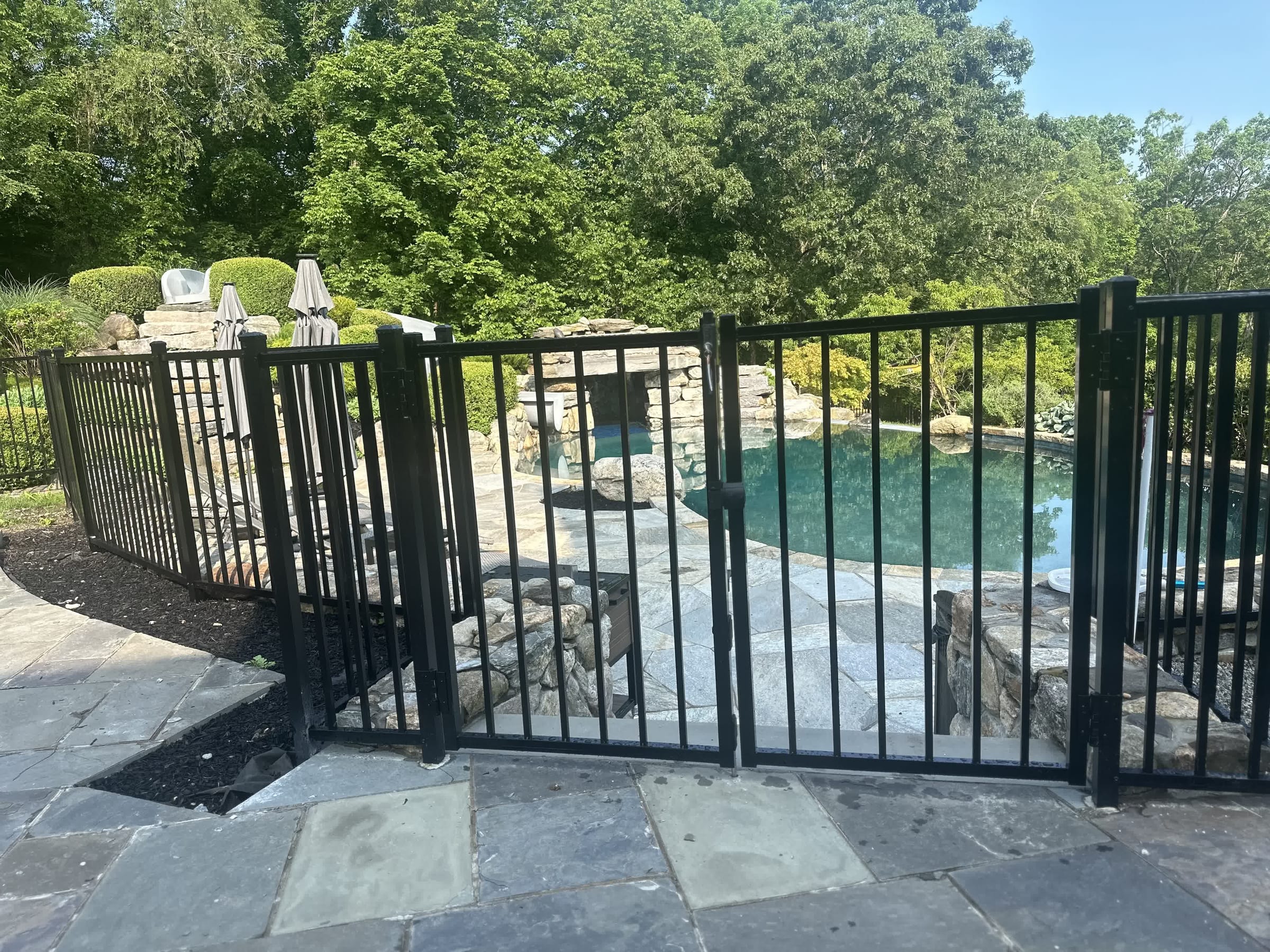 Black aluminum pool safety fence with a self-closing gate around an inground pool on a bluestone patio with stone waterfall