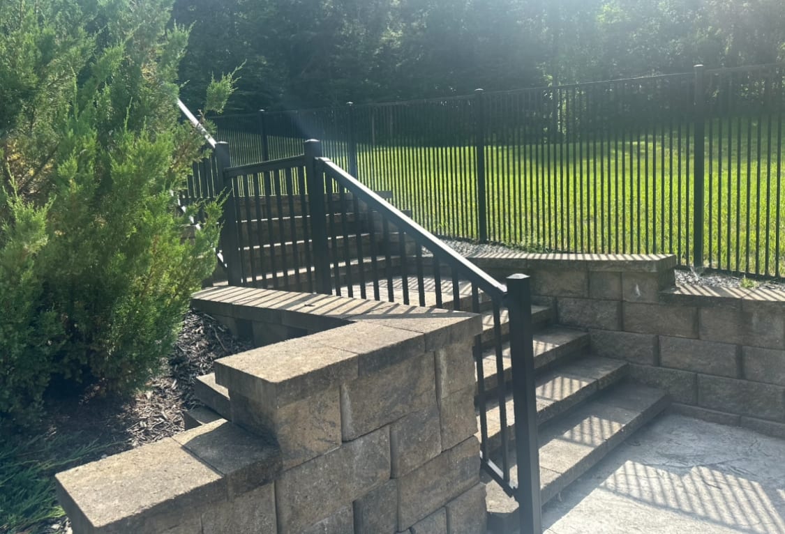 Black aluminum stair railing on paver steps flanked by a stone retaining wall, evergreens and a black yard fence