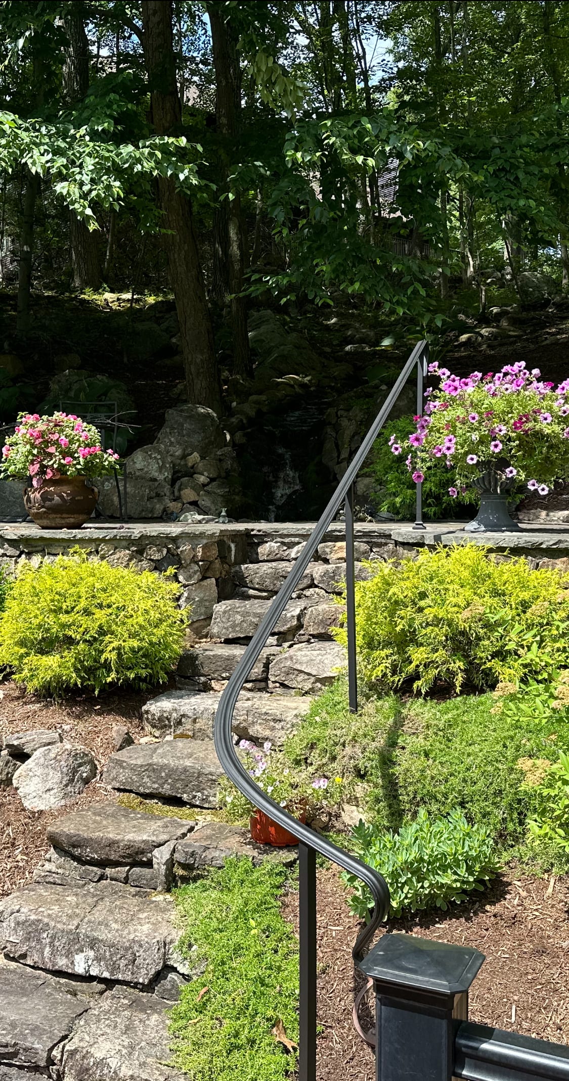 Curved black metal handrail along natural stone steps in a garden with flower urns and a rock water feature