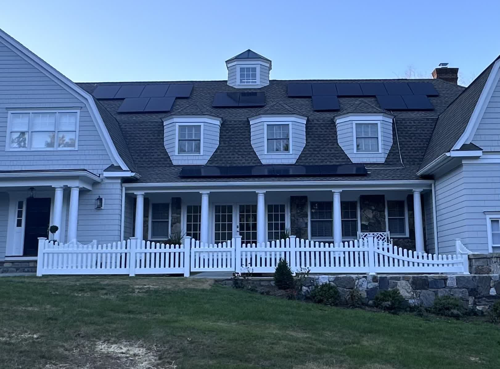 White vinyl scalloped picket fence in front of a large shingled colonial home with stone retaining wall and covered porch.