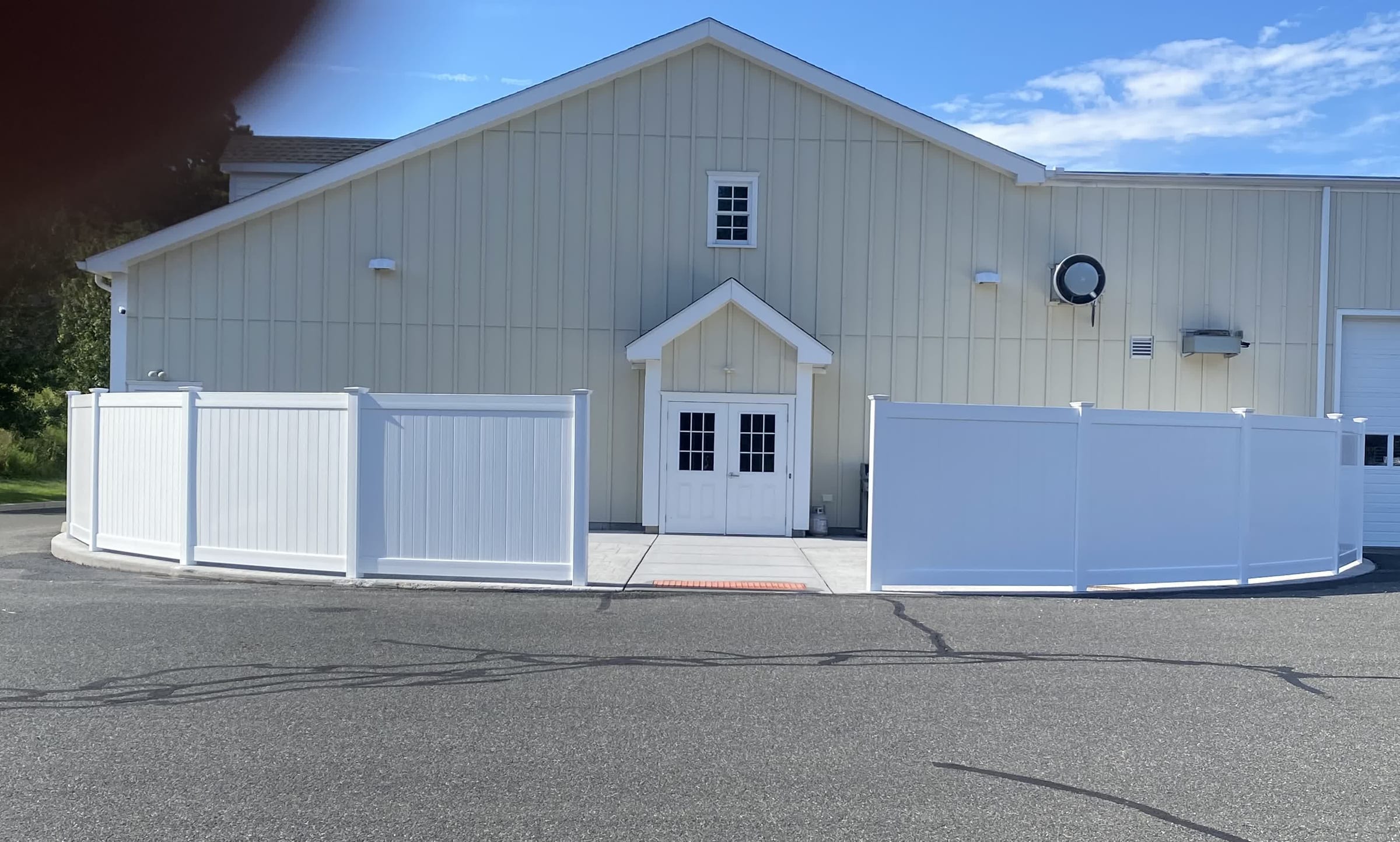 Curved white vinyl privacy fence flanking the entrance of a large commercial barn-style building under blue sky.