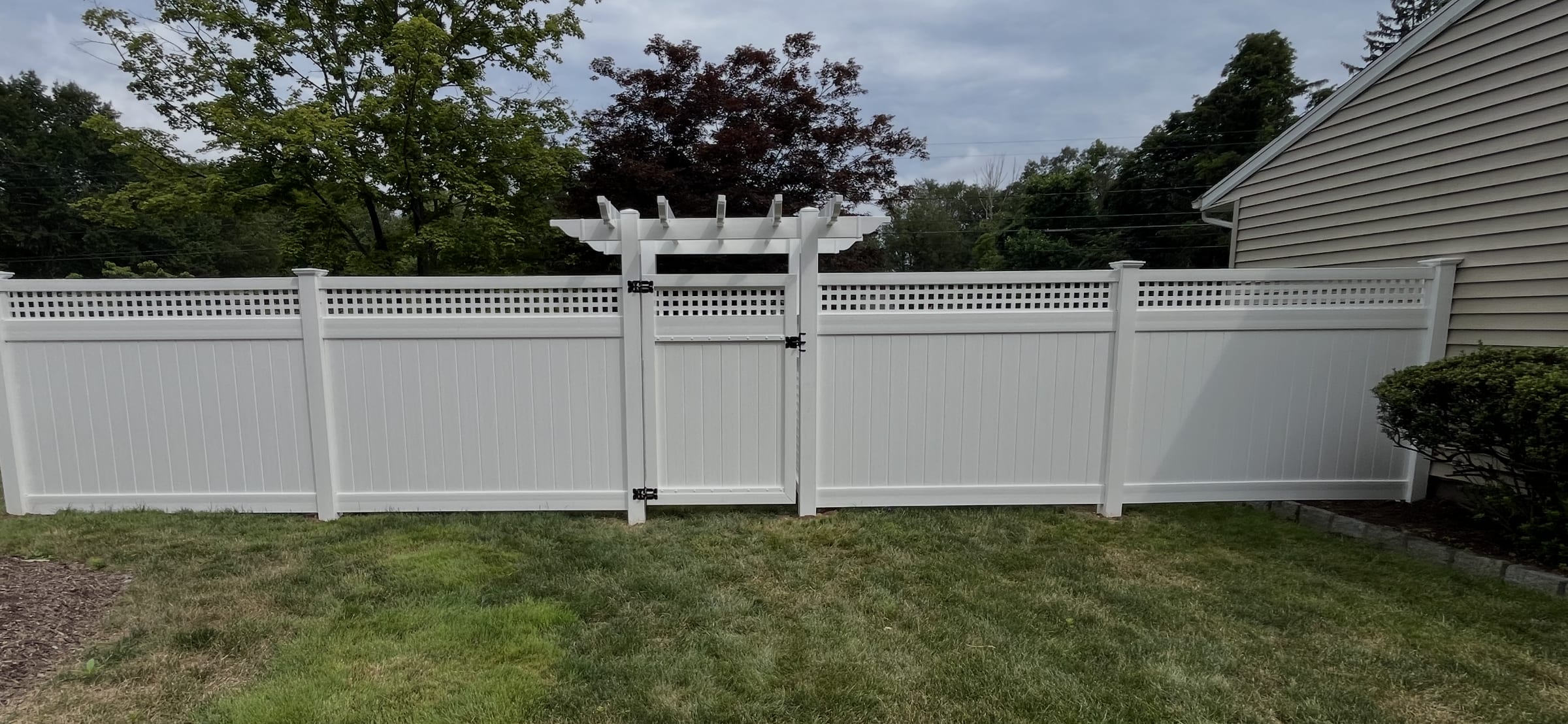 White vinyl privacy fence with square lattice topper, arbor and matching gate along a backyard beside a house.