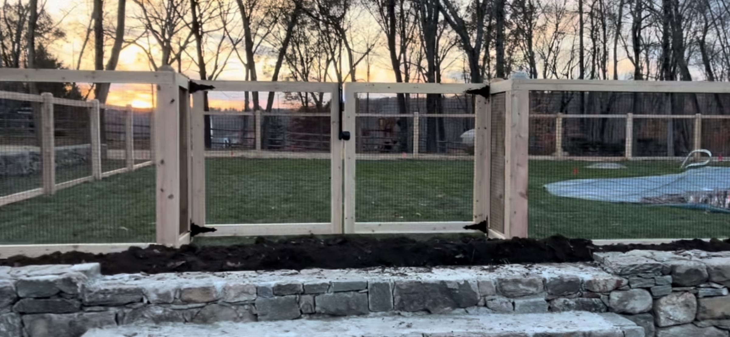 Natural cedar frame fence with black wire mesh and a double gate along a stone retaining wall at dusk