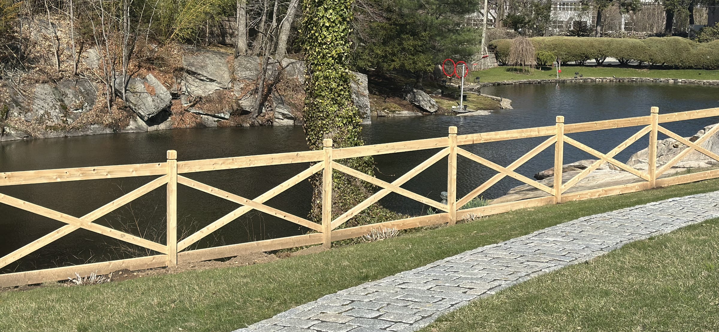 Decorative cedar fence with X-cross panels running beside a rock-lined pond and cobblestone walkway