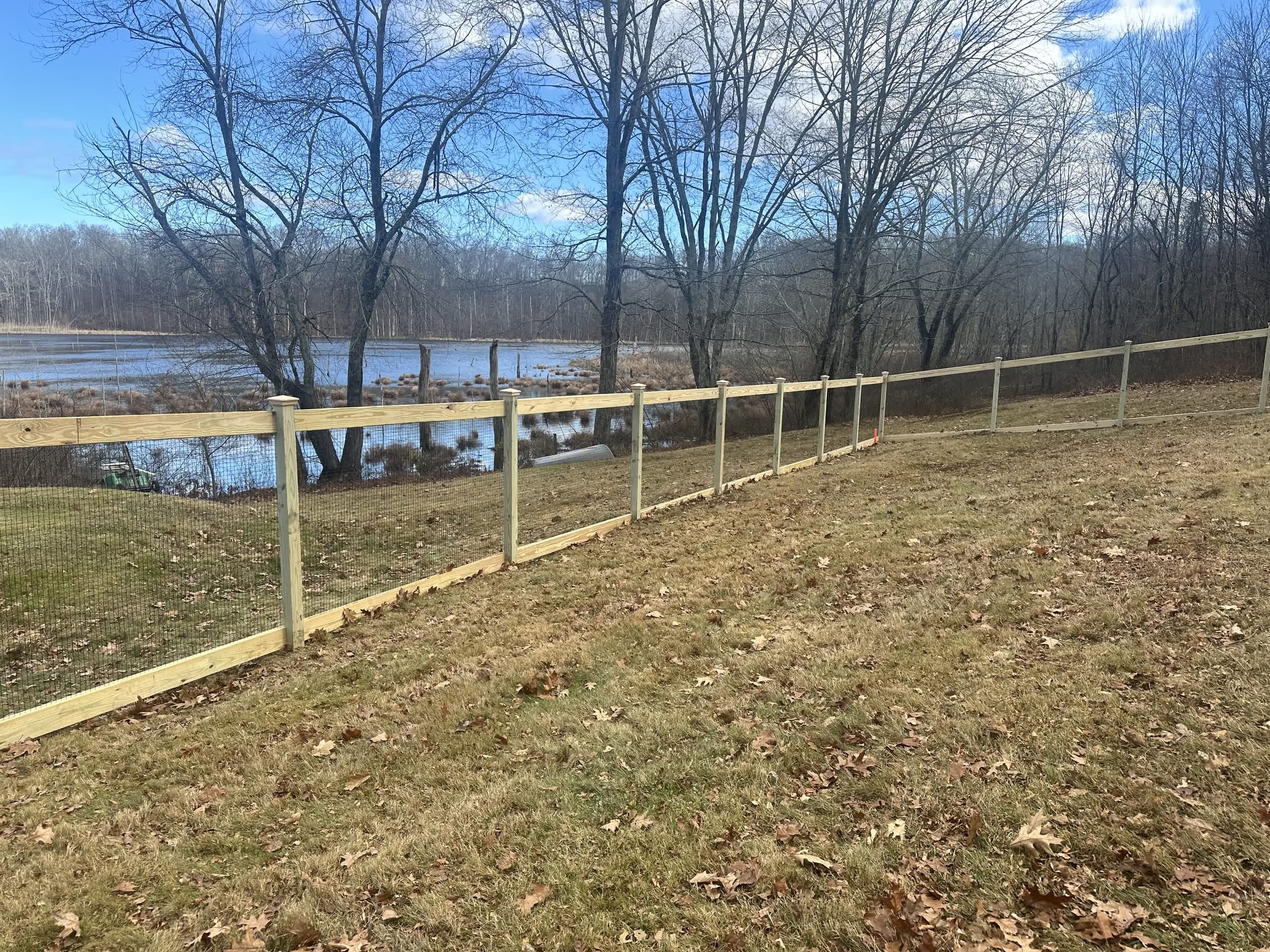 Single-rail pressure-treated wood fence with wire mesh running along a hillside beside a pond